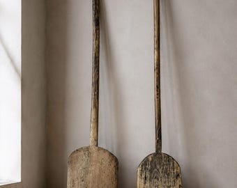 May include: Two tall, weathered wooden peels with long, slender handles and wide, flat, rounded blades. The wood displays a natural, aged texture with visible grain patterns and dark, worn patches, showing signs of extensive use and history.