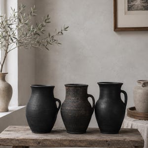 May include: Three dark, matte-finished ceramic pitchers with rounded bodies and single side handles. The two outer pitchers are solid black, while the centre pitcher features a mottled, weathered texture with hints of dark brown and charcoal tones.