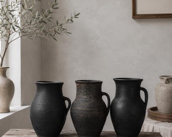May include: Three dark, matte-finished ceramic pitchers with rounded bodies and single side handles. The two outer pitchers are solid black, while the center pitcher features a mottled, weathered texture with hints of dark brown and charcoal tones.