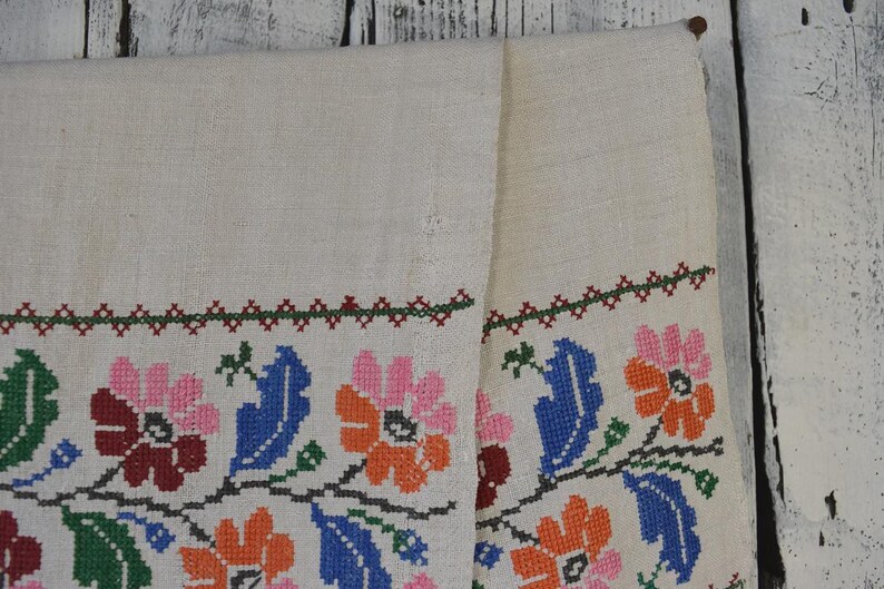 Puede incluir: Un camino de mesa rectangular de color blanco roto con un borde horizontal de bordado en punto de cruz. El patr&oacute;n muestra motivos florales repetidos en tonos rosa, naranja, rojo oscuro, azul y verde, con un borde decorativo.