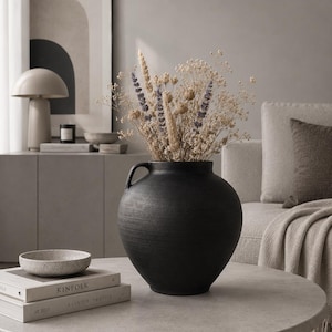 May include: A black, textured ceramic vase with two handles, filled with dried flowers, sits on a round, light gray coffee table. A small, speckled bowl and two books are also on the table. The scene is set in a neutral-toned living room.