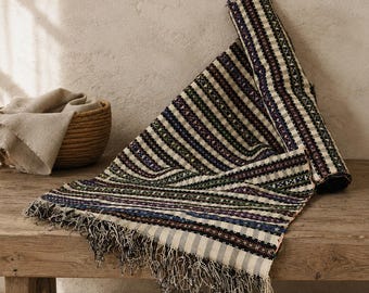May include: A woven textile runner featuring a repeating pattern of vertical stripes in cream, dark blue, and forest green. The fabric has a textured weave and is finished with long, loose fringe along the bottom edge, partially folded over itself.