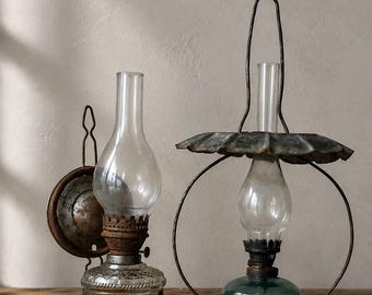 May include: Two vintage oil lamps. The left lamp features a clear, patterned glass base with a rusted metal burner and a tall glass chimney. The right lamp has a green glass base, a metal frame with a circular handle, and a wide, scalloped metal shade.