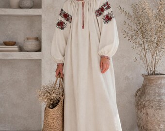 May include: A floor-length cream-colored dress with long, loose sleeves featuring red and green floral embroidery on the shoulders and cuffs. The dress has a gathered round neckline with a thin tie closure and a loose, flowing silhouette.