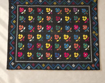 May include: A rectangular black textile featuring a repeating grid of pixelated flowers in shades of pink, yellow, orange, blue, and green. The central floral pattern is enclosed by a white checkered border and a wide outer zigzag frame.