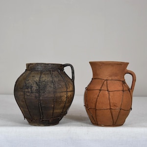 May include: Two antique ceramic jugs. One is dark brown with a wire cage design, the other is terracotta with a similar wire design. Both have handles and are displayed on a white surface.