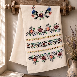 Puede incluir: Una toalla de lino color crema con intrincados bordados de punto de cruz en rojo, azul, verde y amarillo. El diseño incluye motivos florales dispuestos en bandas horizontales y un patrón de corona central, colgada en un perchero de madera.