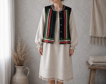 May include: A beige knee-length dress with long sleeves, ruffled cuffs, and a tassel-trimmed hem, worn under a black sleeveless vest. The vest features vertical stripes in red, white, and green, with blue buttons along the front opening.