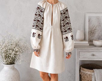 May include: An off-white knee-length dress featuring long, voluminous sleeves with intricate dark red and black floral embroidery. The garment has a round neckline with a central vertical slit and a relaxed, straight-cut silhouette.