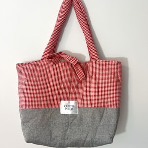 Puede incluir: Un bolso tote a cuadros rojos y blancos con un panel inferior gris y asas a juego. El bolso tiene un lazo en la parte superior y una pequeña etiqueta blanca con el texto "CLOTIS POTIS".