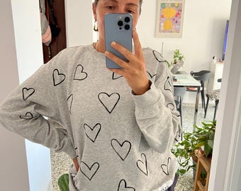 Puede incluir: Una sudadera gris claro con contornos de corazón negros. La sudadera se combina con pantalones a rayas azul marino. Gafas de sol en la cabeza. La imagen es un selfie con espejo.