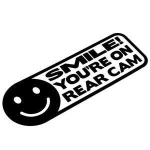 Op de afbeelding: Zwart-witte sticker met een lachend gezicht aan de linkerkant en de tekst "SMILE! YOU'RE ON REAR CAM" in vette letters. De sticker heeft afgeronde hoeken en een rechthoekige vorm.