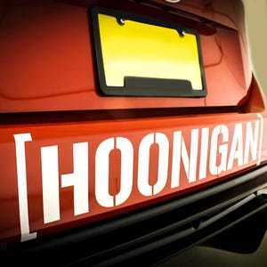 May include: Rear view of a red car featuring a yellow number plate in a black frame. Below, the word "HOONIGAN" is stencilled in white, large capital letters. The car's bumper is black, creating a contrast with the red bodywork and white text.