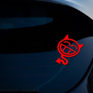 May include: Red devil face sticker with horns, a tail, and a smiling expression. The sticker is attached to the rear window of a dark blue car. The design is a simple, bold graphic.