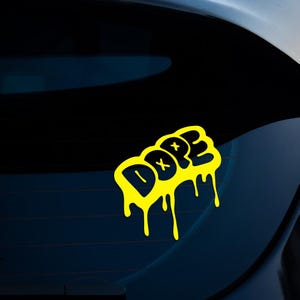 Op de afbeelding: Een gele sticker met het woord "DOPE" in een graffiti-stijl. De letters hebben een druipend effect. De sticker is bevestigd op een donkerblauwe ondergrond, waarschijnlijk een autoruit.