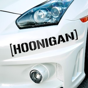 Pode incluir: Um carro branco com um adesivo preto [HOONIGAN] no para-choques dianteiro. O carro tem um para-choques dianteiro preto e uma grelha preta.