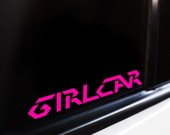 Op de afbeelding: Een felroze vinyl sticker aangebracht op een donker autoraam. De sticker bevat de tekst "GIRLCAR" geschreven in een vet, hoekig en gestileerd geometrisch lettertype. Het ontwerp is scherp, modern en steekt duidelijk af tegen de donkere achtergrond.