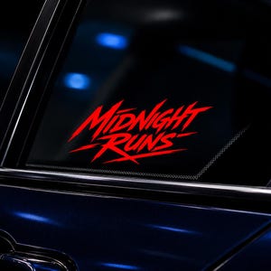 Op de afbeelding: Rode "Midnight Runs" sticker op een autoruit. De tekst is in een gestileerd, hoekig lettertype, wat snelheid en actie suggereert. De sticker is aangebracht op het donker getinte glas van een voertuig, met een glimp van de blauwe lak van de auto.