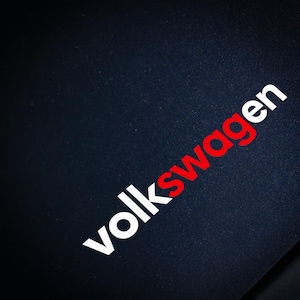 Könnte beinhalten: Auf einer schwarzen Oberfläche steht das Wort "Volkswagen" in weißer und roter Schrift. Das Wort ist diagonal über das Bild geneigt. Der Hintergrund ist eine dunkle, strukturierte Oberfläche.