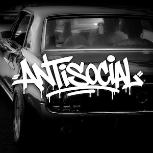 May include: A monochrome image showing the back of a classic car. The word "ANTISOCIAL" is written in white graffiti-style lettering across the car's trunk. The car's windows show blurred figures inside.