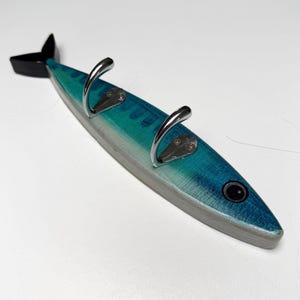 May include: A wall-mounted hook rack shaped like a fish, featuring a blue and teal gradient body with a black tail and eye. Two polished silver-toned metal hooks are attached to the centre of the fish body, each secured with two small screws.