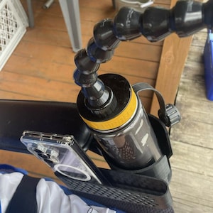 Puede incluir: Una botella de agua negra y amarilla asegurada por un soporte negro, sujeta a un brazo negro flexible. Una funda transparente para teléfono está en un soporte negro debajo de la botella. Los artículos están sobre una superficie de madera.