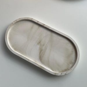 Può includere: Vassoio decorativo ovale con un motivo marmorizzato nei toni del bianco e beige. Il vassoio ha una superficie liscia e lucida e bordi arrotondati. Altri vassoi e oggetti decorativi simili sono sullo sfondo.
