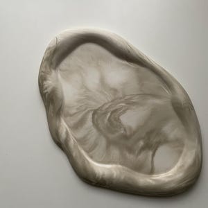 May include: An oval-shaped decorative tray with a marbled design. The tray has a swirling pattern of light grey and white, creating a unique, organic look. The tray is made of a glossy material.