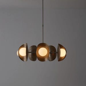 May include: A modern, bronze-toned chandelier with five semi-circular shades, each housing a spherical, glowing light. The fixture hangs from a single rod, creating a balanced and elegant lighting design. The overall aesthetic is contemporary.