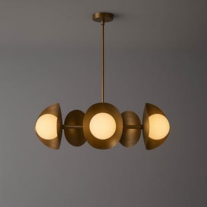 May include: A modern brass chandelier with five light fixtures. The central light is a large, round globe, flanked by two smaller, half-moon shaped lights on each side. The lights have a warm, inviting glow.