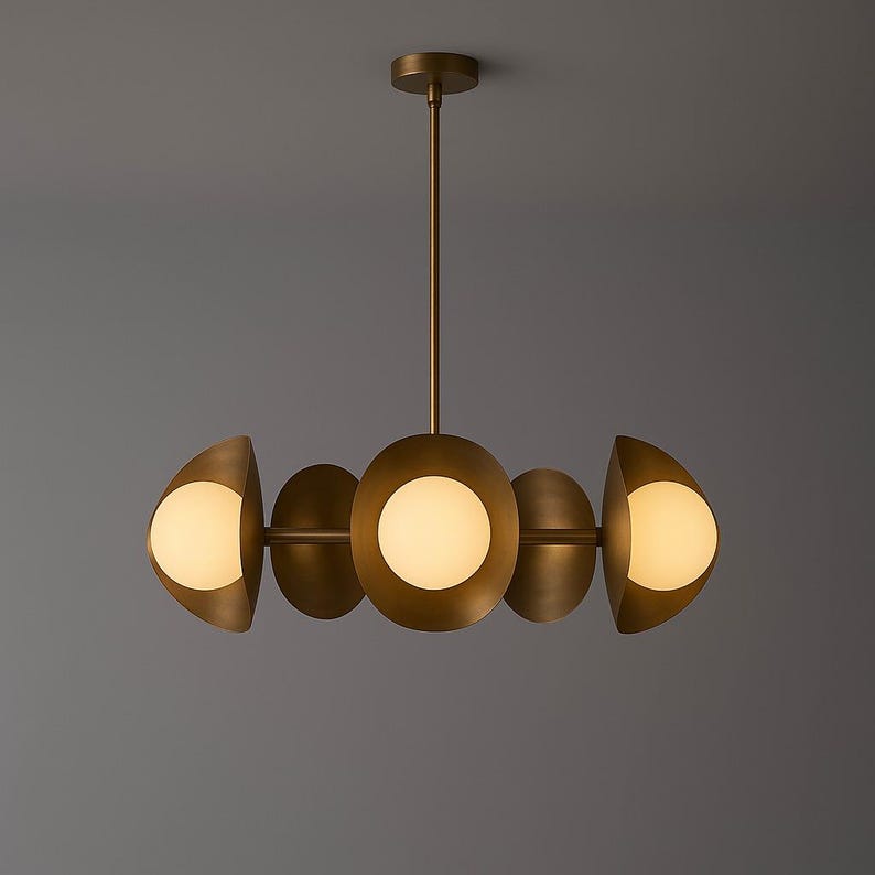 May include: A modern brass chandelier with five light fixtures. The central light is a large, round globe, flanked by two smaller, half-moon shaped lights on each side. The lights have a warm, inviting glow.