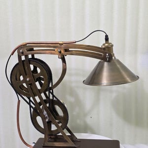 Puede incluir: Lámpara de escritorio de estilo steampunk con acabado bronce. La lámpara presenta un mecanismo de engranajes y cadena, una pantalla cónica y una base de madera marrón oscuro. El diseño incorpora tubos de cobre y un cable de alimentación negro.