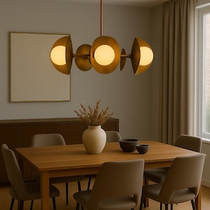 May include: A modern dining room featuring a wooden table and six chairs. Above the table hangs a gold-toned chandelier with spherical lights. A vase with dried plants and two bowls sit on the table. A framed artwork and a cabinet are visible in the background.