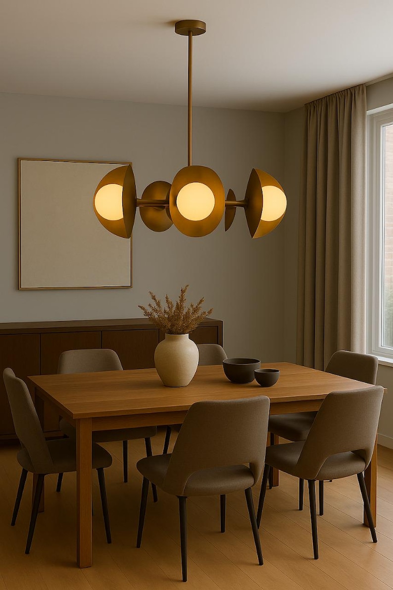 May include: A modern dining room featuring a wooden table and six chairs. Above the table hangs a gold-toned chandelier with spherical lights. A vase with dried plants and two bowls sit on the table. A framed artwork and a cabinet are visible in the background.