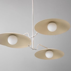 May include: A modern, three-light chandelier with a minimalist design. The fixture features three beige, saucer-shaped shades, each housing a spherical white light. The metal frame is silver, and the overall aesthetic is clean and contemporary.