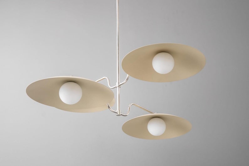 May include: A modern, three-light chandelier with a minimalist design. The fixture features three beige, saucer-shaped shades, each housing a spherical white light. The metal frame is silver, and the overall aesthetic is clean and contemporary.