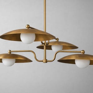 May include: A brass-toned chandelier with four downward-facing, saucer-shaped shades. Each shade holds a spherical, white light. The fixture has a central stem and curved arms, creating a modern, elegant design.