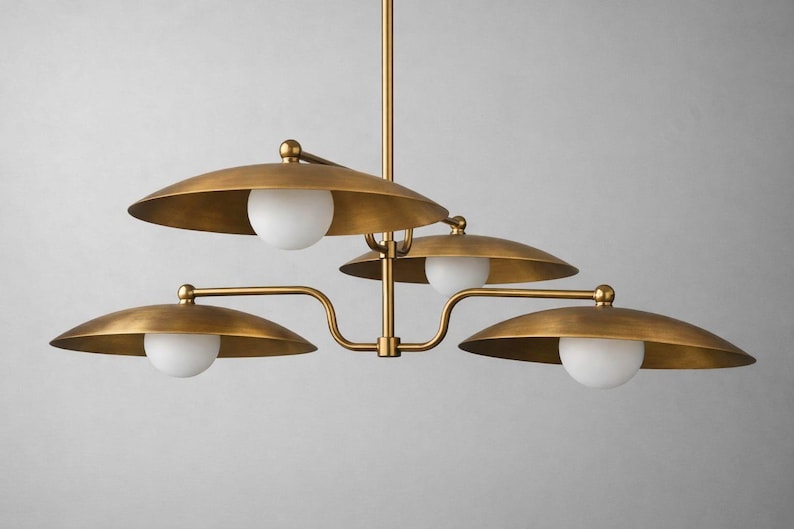 May include: A brass-toned chandelier with four downward-facing, saucer-shaped shades. Each shade holds a spherical, white light. The fixture has a central stem and curved arms, creating a modern, elegant design.