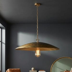 Op de afbeelding: Een grote, antieke messing hanglamp met een brede, ondiepe kap. Het licht hangt aan een ketting en heeft een Edison-lamp in vintage-stijl. De lamp hangt in een kamer met donkere muren.