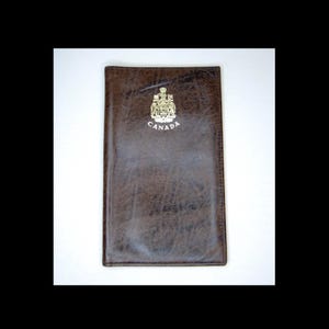 Puede incluir: Una cartera de cuero marrón con la palabra "CANADA" en letras doradas. La cartera tiene un emblema dorado en la parte superior. La cartera es rectangular y tiene una superficie texturizada.