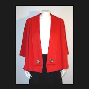 Puede incluir: Chaqueta roja brillante estilo capa, con cuello ancho y mangas cortas. La chaqueta tiene dos cierres de botón plateados. Se muestra en un maniquí, combinada con una camiseta blanca y una falda negra.