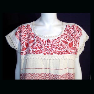 Puede incluir: Top blanco bordado con diseños florales y de pájaros rojos. La blusa de manga corta presenta intrincados bordados rojos alrededor del cuello y las mangas, con un patrón detallado en el pecho. El dobladillo inferior tiene un diseño geométrico rojo.