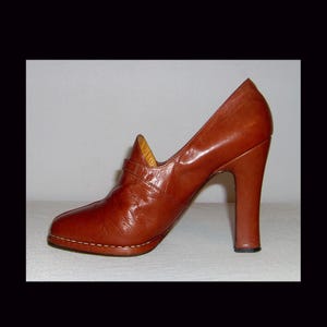 Può includere: Una décolleté in pelle marrone con tacco alto a blocco. La scarpa presenta una punta arrotondata, un dettaglio con cinturino sul collo del piede e una suola cucita. Il tacco misura circa 7,6 cm.