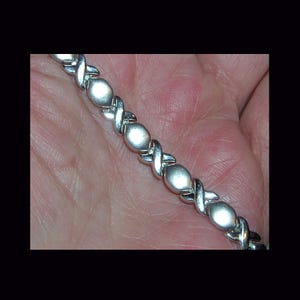 Puede incluir: Una pulsera plateada con un patrón alterno de formas X y O. La pulsera tiene un acabado pulido y se muestra sobre un fondo color piel. La pulsera es una joya.