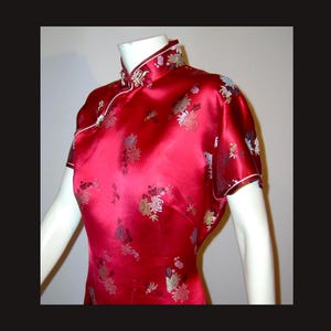 Puede incluir: Vestido de satén rojo con mangas cortas y cuello mandarín. El vestido presenta un estampado floral en tonos dorados, plateados y morados. Ribetes blancos acentúan el cuello y las mangas. El vestido se exhibe en un maniquí.