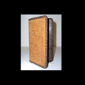 Puede incluir: Una cartera de cuero color canela con un diseño floral y costuras marrones. La cartera tiene un patrón en relieve de flores y hojas. Los bordes están cosidos con hilo marrón. Se ve una sección marrón oscuro en el lateral.