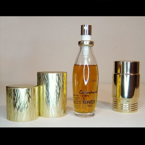 May include: A gold and clear glass bottle of Cabochard perfume by Gres Paris. The bottle is sitting on a gold metal stand with a round top and three legs. The bottle has a gold cap with a silver spray nozzle.