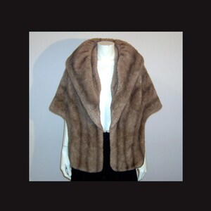 May include: A brown faux fur cape with a wide collar and short sleeves. The cape is displayed on a mannequin, showcasing its soft texture and elegant design. The garment is a neutral colour, suitable for various outfits.