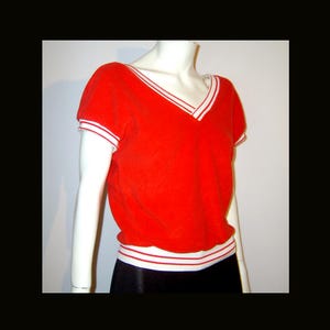 Puede incluir: Una camiseta roja brillante con cuello en V y ribetes a rayas blancas y rojas en el escote, las mangas y la cintura. La camiseta de manga corta se muestra en un maniquí.