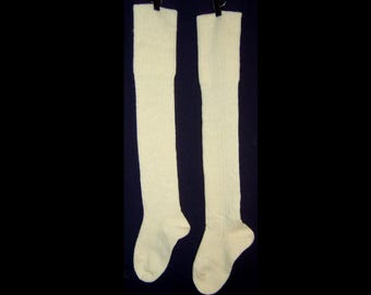 May include: Two off-white, knee-high socks hanging against a black background. The socks are made of a ribbed knit material and are suspended by black clips. The socks are a solid color and appear to be new.